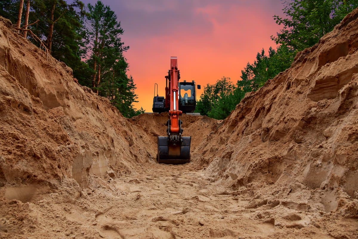 Excavator digging a trench for mainline sewer pipe replacement at sunset